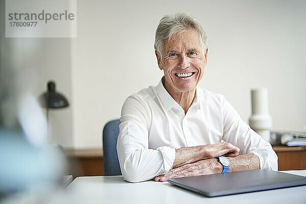 Glücklicher älterer Geschäftsmann mit Laptop am Schreibtisch im Studio