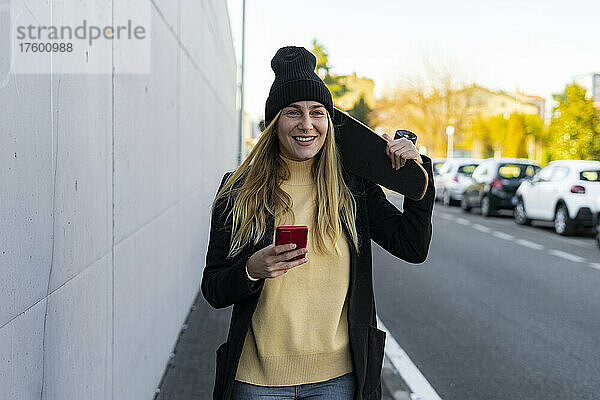 Blonde Frau mit Smartphone und Skateboard an der Wand auf der Straße