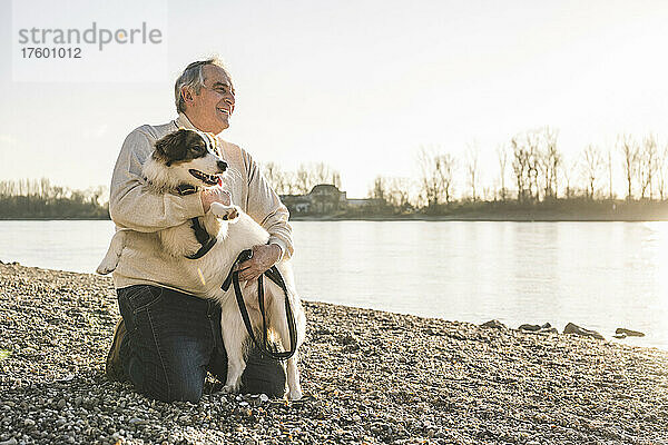 Glücklicher älterer Mann umarmt Hund am Strand an einem sonnigen Tag