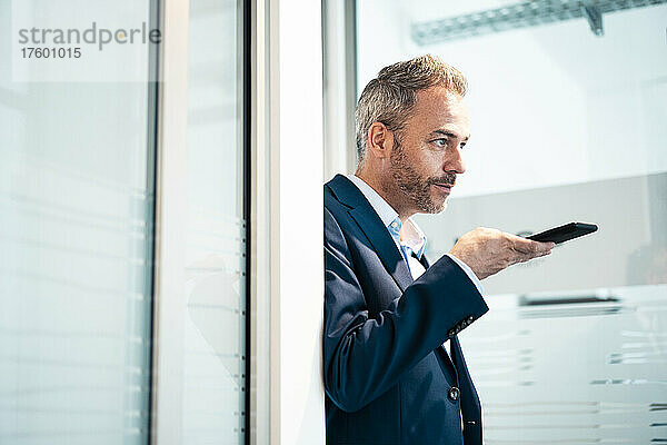 Businessman talking through smart phone speaker leaning on doorway at office