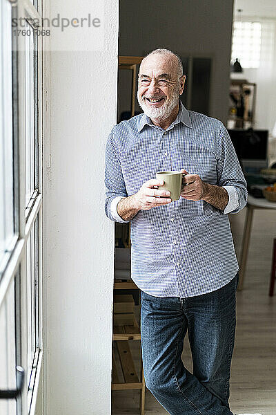 Glücklicher Geschäftsmann hält eine Kaffeetasse in der Hand und lehnt zu Hause an der Wand