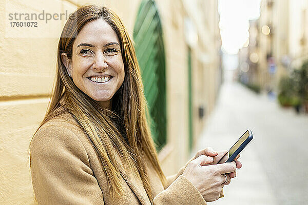 Glückliche Frau mit Mobiltelefon  die an der Wand steht