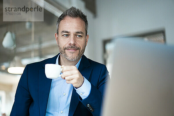 Businessman holding coffee cup looking at laptop in office