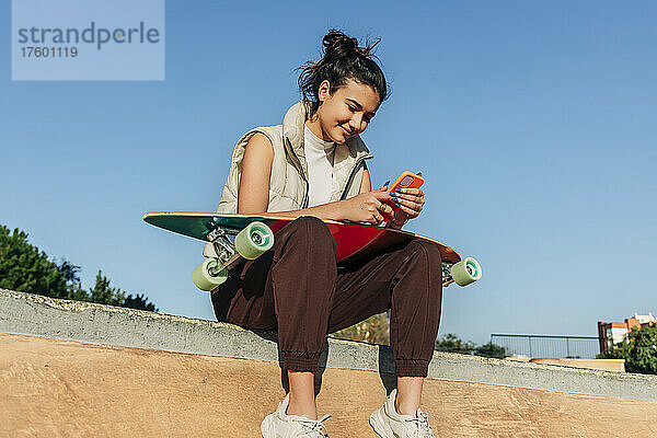 Frau mit Skateboard benutzt Mobiltelefon auf Sportrampe im Park