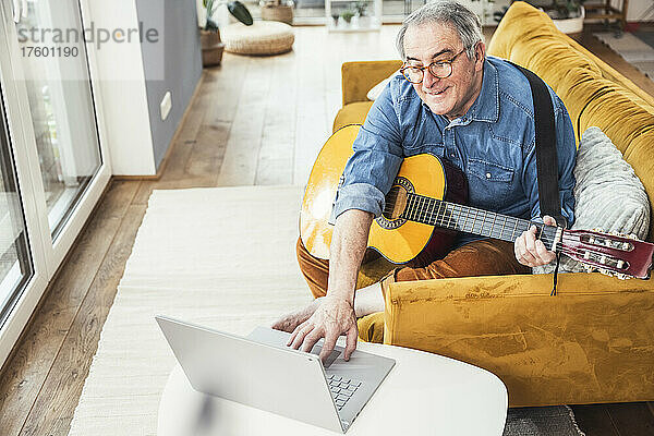 Älterer Mann mit Gitarre schaut sich Online-Tutorial auf Laptop im Wohnzimmer an