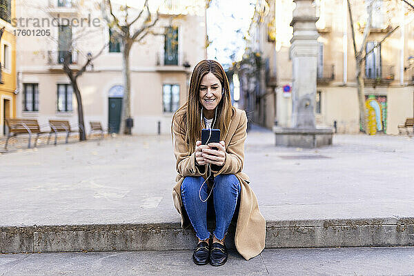 Lächelnde Frau  die auf dem Fußweg sitzt und mit ihrem Mobiltelefon im Internet surft