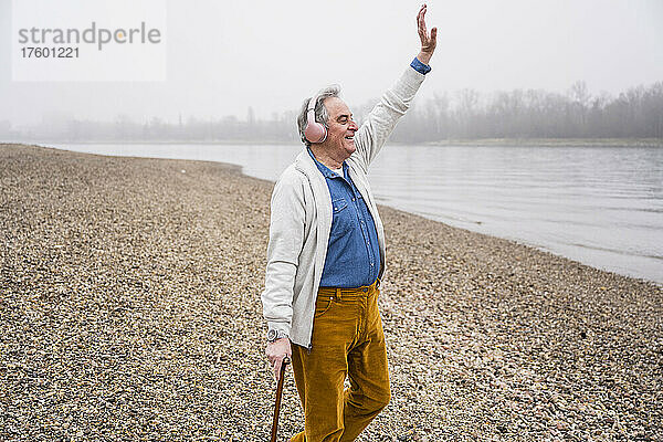 Glücklicher älterer Mann mit erhobener Hand  der am Strand Musik über Kopfhörer hört