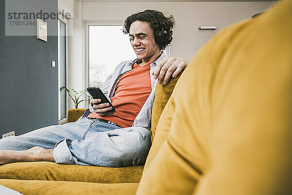 Fröhlicher Mann  der im Wohnzimmer Musik über kabellose Kopfhörer hört und sein Smartphone nutzt