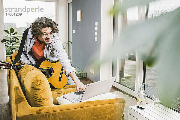 Lächelnder junger Mann mit Gitarre und Laptop auf dem heimischen Sofa