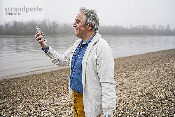 Lächelnder älterer Mann  der am Strand einen Videoanruf über sein Mobiltelefon tätigt