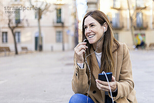 Glückliche Frau mit braunen Haaren  die über In-Ear-Kopfhörer mit ihrem Smartphone spricht