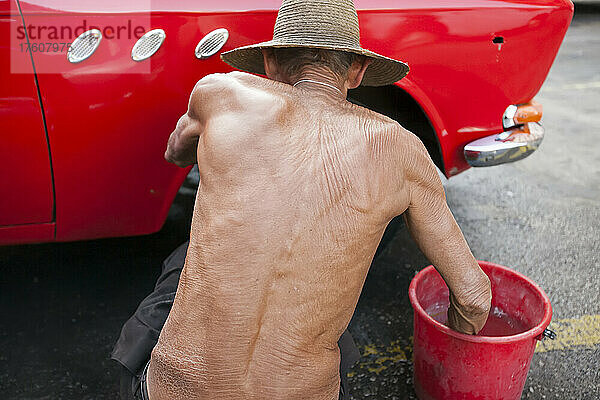 Ein älterer Mann greift in einen Wassereimer  während er einen amerikanischen Oldtimer von Hand wäscht; Havanna  Kuba
