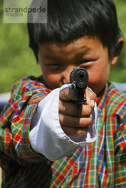 Ein Junge in traditioneller Kleidung richtet ein Spielzeuggewehr auf die Kamera; Paro  Bhutan