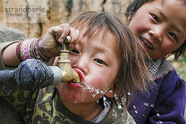 In einem Dorf in Phobjika trinkt ein junges Mädchen Wasser aus einem Zapfhahn  während ein anderes Mädchen zusieht; Phobjika  Bhutan