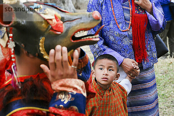 Ein kleiner Junge schaut zu einem Mann auf  der eine Maske trägt  um beim bhutanischen Paro Tshechu Festival im Paro Dzong  einem Kloster und einer Festung in Paro  Bhutan  einen Cham-Tanz aufzuführen; Paro  Bhutan