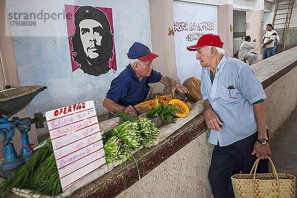 Zwei ältere Männer  einer davon ein Obst- und Gemüsehändler  unterhalten sich auf einem Bauernmarkt in Cienfuegos  Kuba. Ein Gemälde von Che Guevara ziert die Wand; Cienfuegos  Kuba