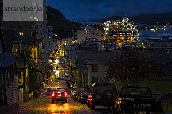 Die Bremslichter eines Autos erhellen die dunkle Straße an einem regnerischen Abend in Juneau. Angedockte Kreuzfahrtschiffe erhellen in der Abenddämmerung den Hafen  der wie ein Gebäude in der Stadt aussieht. Der Tourismus ist ein wachsender Wirtschaftszweig in Alaskas Südosten; Juneau  Alaska  Vereinigte Staaten von Amerika