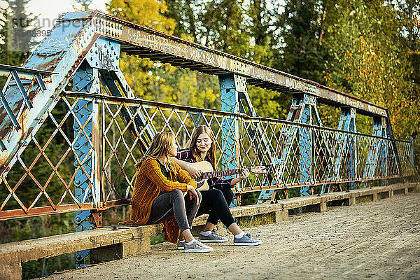 Zwei Schwestern sitzen auf einer Parkbrücke  eine mit einer Akustikgitarre  und genießen gemeinsam die Musik; Edmonton  Alberta  Kanada