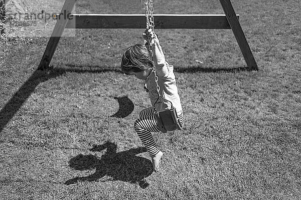 Ein junges Mädchen sitzt auf einer Schaukel und schaut auf ihren Schatten  der auf den Boden geworfen wird; North Vancouver  British Columbia  Kanada