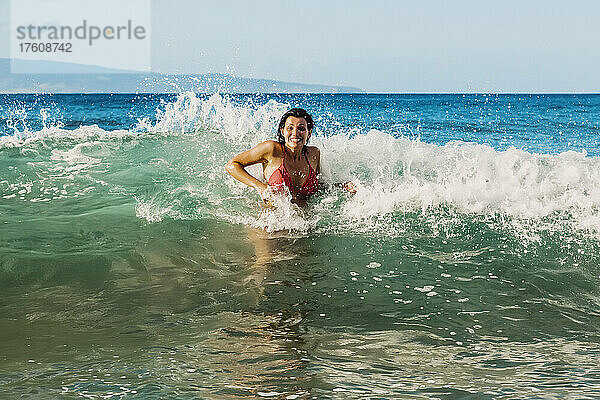 Frau in einem roten Bikini steht im Wasser und wird von einer Welle am D. T. Fleming Beach mit der Insel Molokai in der Ferne bespritzt; Maui  Hawaii  Vereinigte Staaten von Amerika