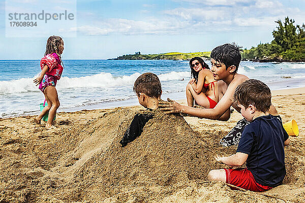 Mutter sitzt am D. T. Fleming Beach am Rande des Wassers und beobachtet ihre Kinder  die spielen und ein Geschwisterchen im Sand vergraben; Kapalua  Maui  Hawaii  Vereinigte Staaten von Amerika