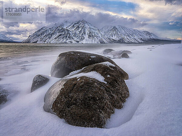 Dramatische Winterlandschaft mit eisbedeckten Felsen am zugefrorenen Ufer des Kathleen Lake und den Wolken über dem Mount Worthington in der Ferne  die eine stimmungsvolle Atmosphäre schaffen; Haines Junction  Yukon  Kanada