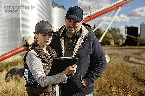 Älteres Ehepaar bei der Arbeit auf dem Bauernhof  vor Getreidesilos und einer Schnecke stehend und ihren Tablet-Computer konsultierend; Alcomdale  Alberta  Kanada