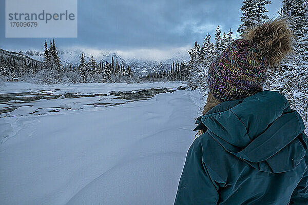 Rückenansicht einer Frau  die im Schnee steht und die Aussicht entlang des Wheaton River im Winter mit den Bergen in der Ferne genießt; Whitehorse  Yukon  Kanada