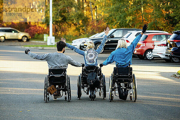 Drei junge Querschnittsgelähmte in ihren Rollstühlen  die an einem schönen Herbsttag mit erhobenen Armen in einer Reihe über einen Parkplatz in einem Park fahren; Edmonton  Alberta  Kanada
