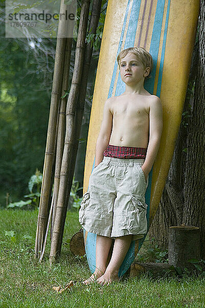 Ein sechsjähriger Junge mit hochgezogenen Boxershorts lehnt sich an ein Surfbrett; Cabin John  Maryland.
