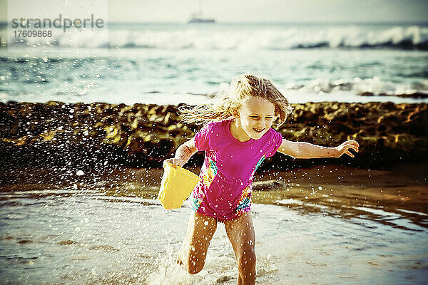 Ein junges Mädchen läuft mit einem Eimer in der Hand vom Meer ans Ufer  während das Wasser auf sie spritzt; Ka'anapali  Maui  Hawaii  Vereinigte Staaten von Amerika