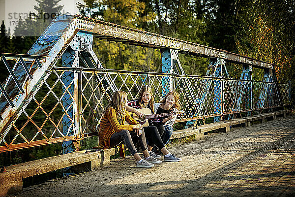 Mutter und zwei Schwestern sitzen auf einer Parkbrücke  eine mit einer Akustikgitarre  und genießen gemeinsam die Musik; Edmonton  Alberta  Kanada