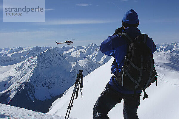 Ein Skifahrer beobachtet  wie sich ein Heli-Ski-Helikopter einem Gipfel nähert; Selkirk Mountains  British Columbia  Kanada.