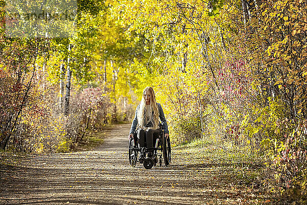 Junge querschnittsgelähmte Frau in ihrem Rollstuhl  die an einem schönen Herbsttag einen Weg in einem Park hinunterfährt; Edmonton  Alberta  Kanada