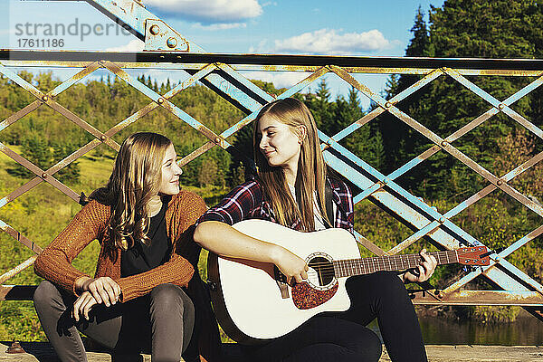 Zwei Schwestern sitzen auf einer Parkbrücke  eine mit einer Akustikgitarre  und genießen gemeinsam die Musik; Edmonton  Alberta  Kanada