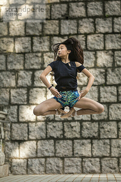 Mädchen tanzt mit Ballettschuhen und in einer Pose in der Luft gegen eine Steinmauer in einem öffentlichen Park; Hongkong  China