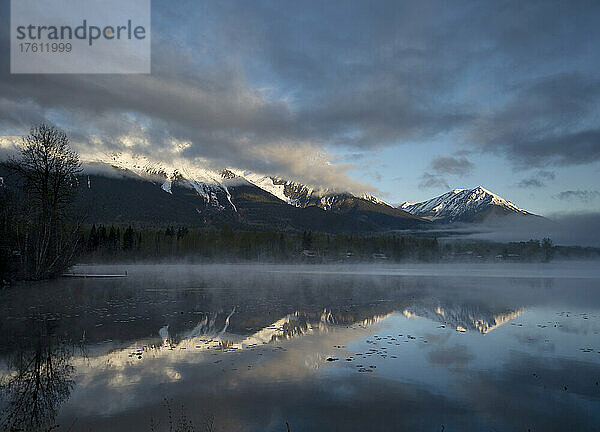 Der schroffe Gipfel des Hudson Bay Mountain ist mit Schnee bedeckt  im Vordergrund steigt bei Sonnenaufgang Nebel vom See auf; Smithers  British Columbia  Kanada