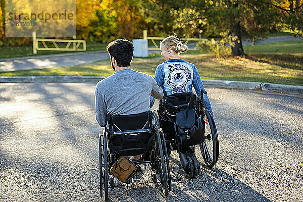 Zwei junge Querschnittsgelähmte in ihren Rollstühlen fahren an einem schönen Herbsttag über einen Parkplatz in einem Park; Edmonton  Alberta  Kanada
