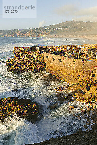 Wellen des Atlantischen Ozeans am Ufer des Praia do Guincho in der Nähe von Cascais mit dem Außenschwimmbad des Hotels Estalagem Muchaox  das innerhalb der Steinmauern einer alten Festung gebaut wurde; Praia do Guincho  Cascais  Lison  Portugal