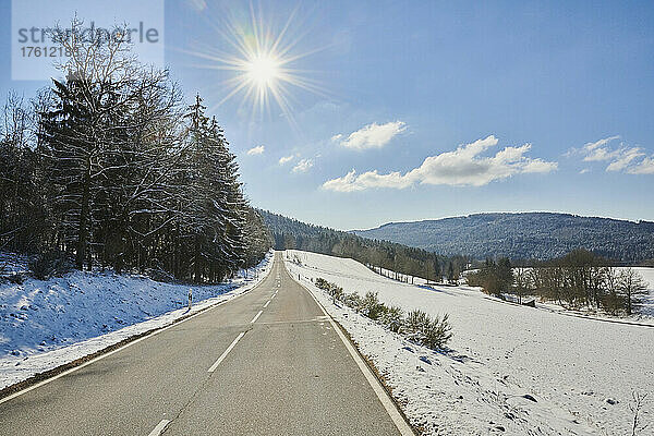 Zweispurige asphaltierte Straße durch eine verschneite Landschaft mit einem Sonnenaufgang am blauen Himmel; Bayern  Deutschland