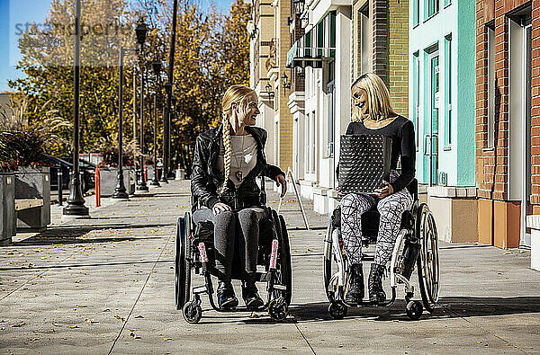 Zwei junge  querschnittsgelähmte Frauen in ihren Rollstühlen  die an einem schönen Herbsttag gemeinsam auf einem Gehweg spazieren gehen  eine hält eine Einkaufstasche; Edmonton  Alberta  Kanada