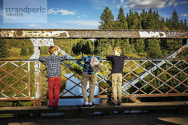 Drei Jungen blicken von einer Parkbrücke auf einen darunter liegenden Bach  ein Junge in der Mitte beugt sich nach hinten  um in einer albernen Pose in die Kamera zu schauen; Edmonton  Alberta  Kanada