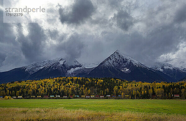 Weite und dramatische Landschaft mit schroffen Berggipfeln unter einem bewölkten Himmel und einem Zug  der an einem herbstlich gefärbten Wald vorbeifährt; British Columbia  Kanada