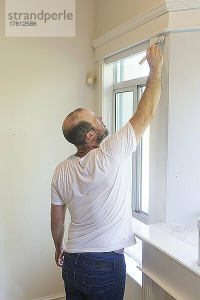 Ein Mann streicht zu Hause eine Wand und eine Verkleidung mit einem Pinsel; North Vancouver  British Columbia  Kanada
