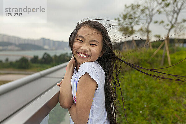 Mädchen mit verwehtem Haar lächelt  während sie sich die Haare aus dem Gesicht hält; Hongkong  China