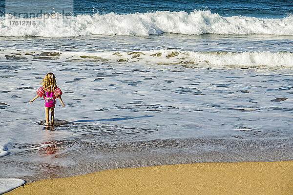 Blick von hinten auf ein junges Mädchen  das vom Strand ins Wasser geht  während die Wellen an das Ufer von D.T. Fleming rollen; Kapalua  Maui  Hawaii  Vereinigte Staaten von Amerika
