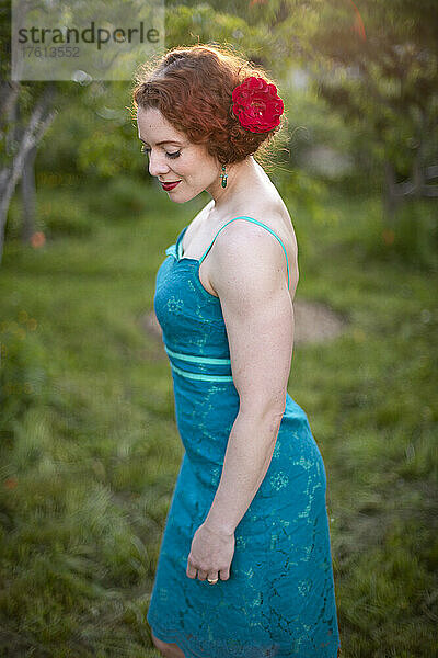 Porträt einer Frau mit roten Haaren steht in einem formellen ärmellosen Kleid mit einer Blume im Haar in einem Garten bei Sonnenuntergang; British Columbia  Kanada