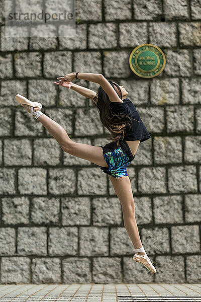 Mädchen tanzt mit Ballettschuhen und in einer Pose in der Luft gegen eine Steinmauer in einem öffentlichen Park; Hongkong  China