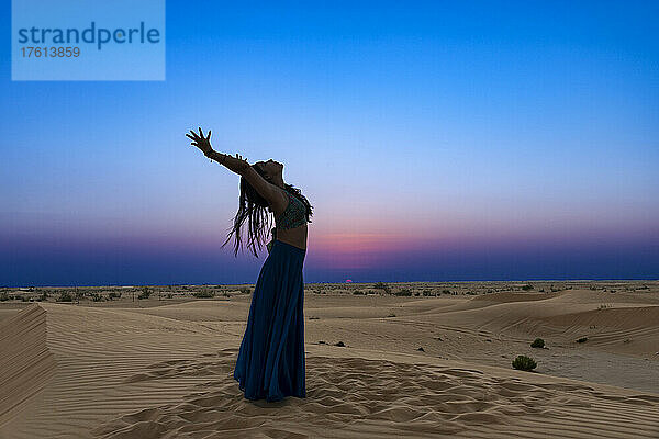 Frau mit langem Rock im Gegenlicht des Sonnenuntergangs auf dem Wüstensand  stehend in einer Rückbeuge-Yoga-Pose; Abu Dhabi  Vereinigte Arabische Emirate