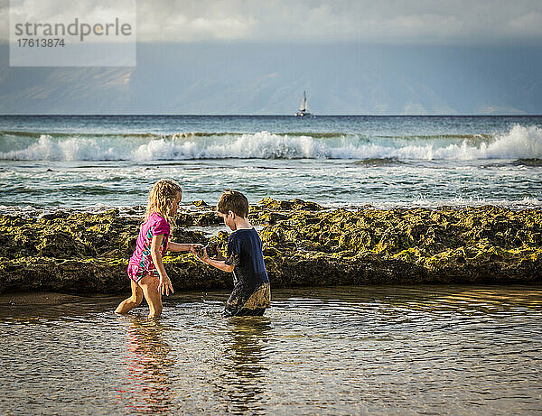 Ein Junge und ein Mädchen untersuchen einen Felsen  während sie am Strand von Ka'anapali im Meer stehen  mit Blick auf die Insel Molokai und ein Segelboot in der Ferne; Ka'anapali  Maui  Hawaii  Vereinigte Staaten von Amerika